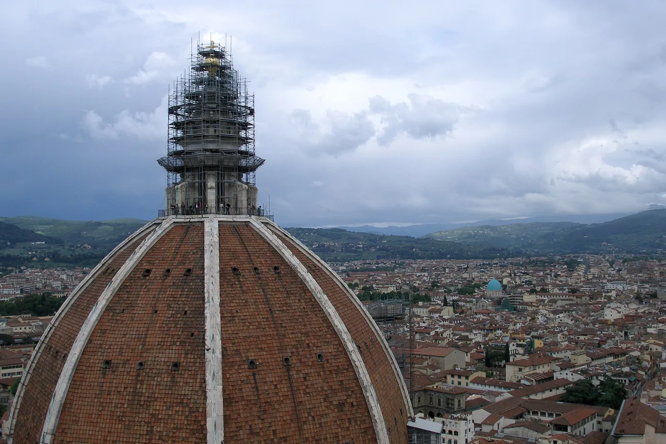 Brunelleschi's dome seen from above and to one side: red tile panels separated by white marble ribs rising to the lantern, which is wrapped in scaffolding, with the rooftops of Florence and green hills beyond under a cloudy sky.