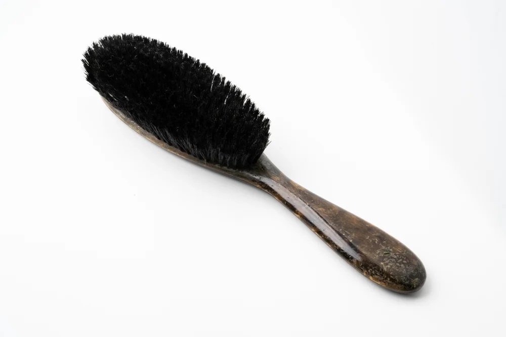 A clothes brush with a dense oval head of black bristle and a long tapered wooden handle with worn, stained finish.