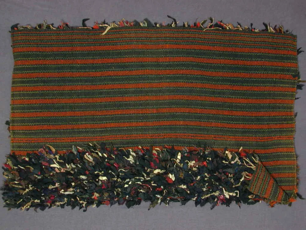 A Norwegian boat rug lying folded back: the smooth side is woven in narrow red and dark green stripes, and one end shows the shaggy pile of knotted rags and wool in dark blue, red, and cream.