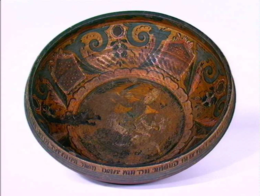 Inside a large old wooden ale bowl: dark green C-scrolls and small flowers on an ochre ground, painted panels, a central painted medallion, and a lettered inscription around the rim.
