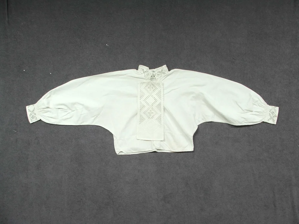 A white cotton bunad shirt laid flat, with a band of Hardanger cutwork on the stand collar, on both cuffs, and on a long rectangular front bib in a diamond lattice.