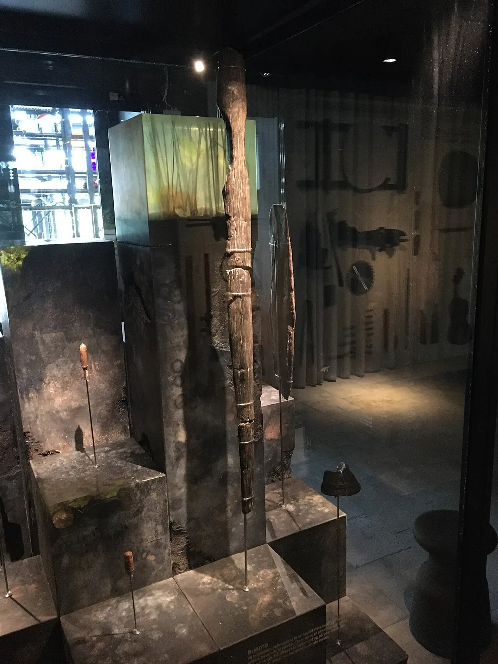 In a dim museum case, a dark, weathered wooden bow limb stands upright, bound with thin cords, beside a second narrow limb fragment and small arrow pieces on pins.