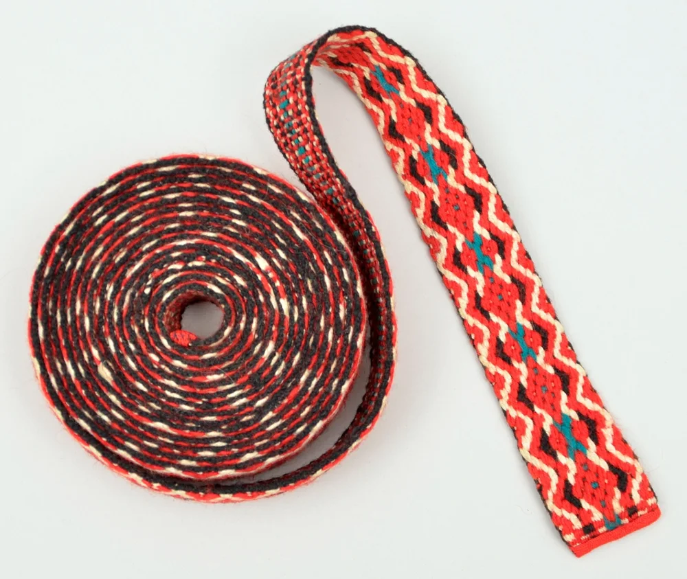 A long woven band, mostly coiled, with one end unrolled to show a red, black, and white zigzag pattern with small teal accents down the center and a red-bound end.