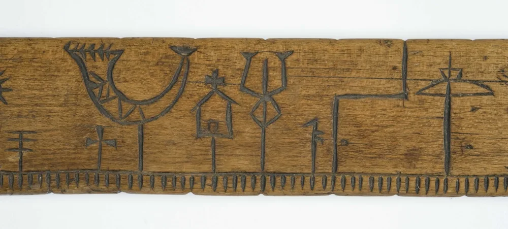 Close-up of a carved wooden primstav: a row of short notches along the bottom edge, one per day, and stick-figure symbols rising from them: a large horn shape, small crosses, a little house with a cross, a three-pronged figure, and a wide cross shape.