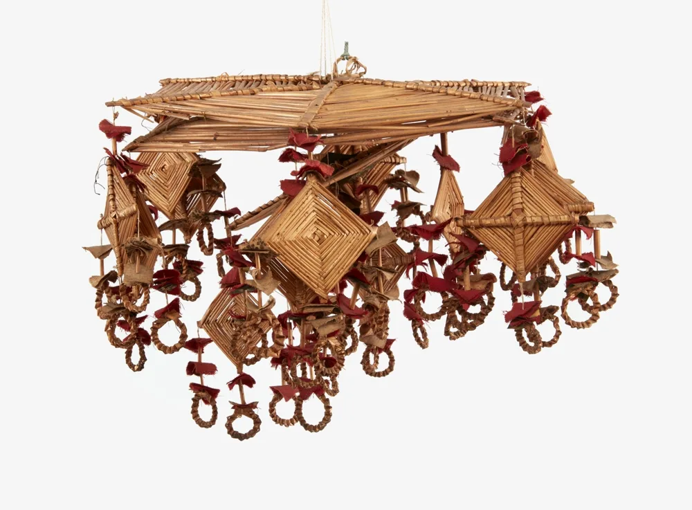A straw crown with a large square roof of straws and a smaller roof beneath it; from its corners hang large straw diamonds of concentric squares on crossed straws, strings of straw pieces with red cloth patches between them, and twisted straw rings at the bottom.