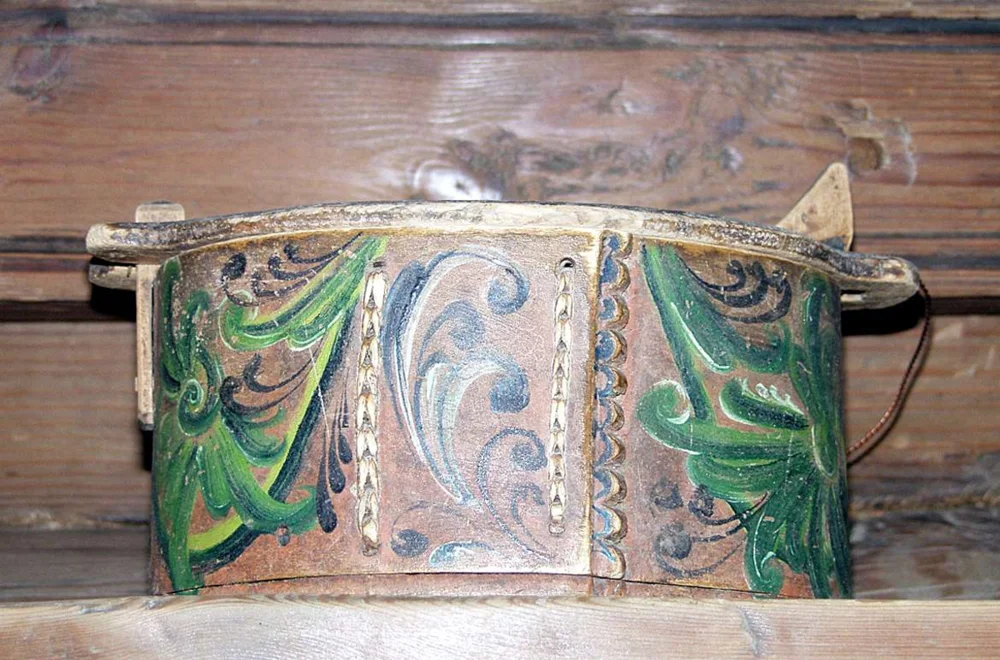 An oval pine tine painted with green, blue, and white rosemaling on brown, with carved scalloped lap edges and stitched seams; the lid's ends pass between upright posts at the sides, and a cord hangs at one end.