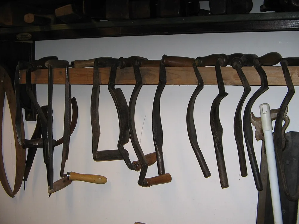 A row of old coopers' knives hangs from a wooden rail: drawknives with straight and curved blades, deep hook-shaped hollowing knives, and a small saw-toothed tool, with a wooden hoop at the left.