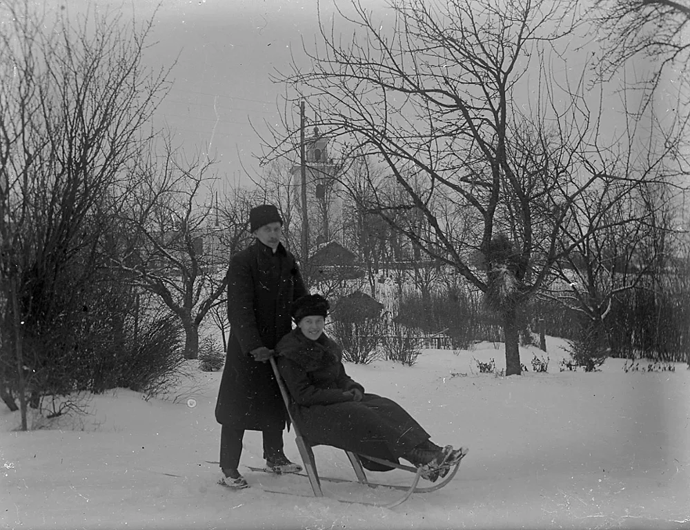 An old black-and-white photograph of a snowy yard with bare trees: a man in a long dark coat and hat stands behind a kick sled holding the handlebar, while a woman in a dark coat sits on its seat.