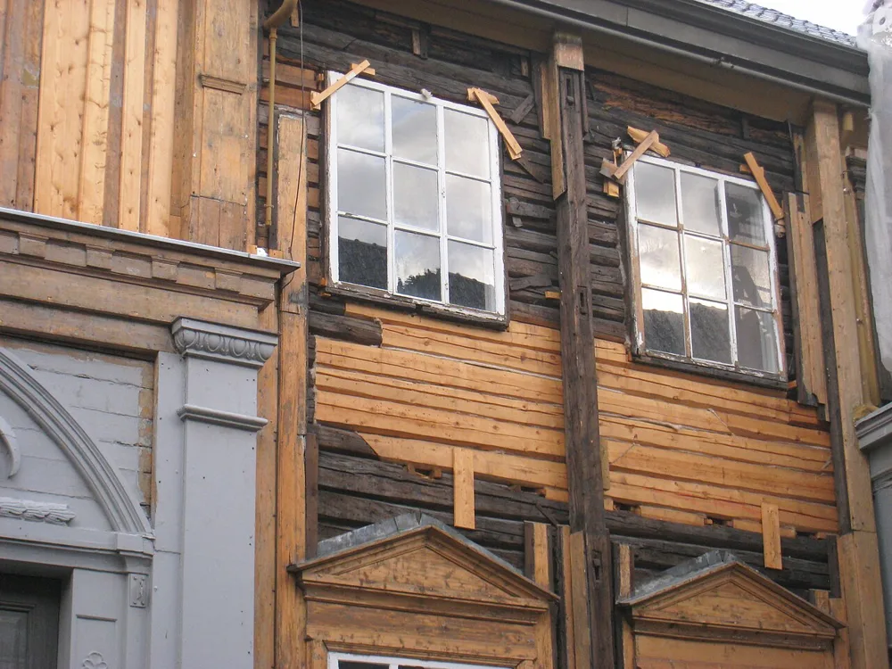 Two old multi-pane windows in a log wall exposed during restoration at Stiftsgården in Trondheim, with the cladding stripped away around them and new boards patched into the timber below.