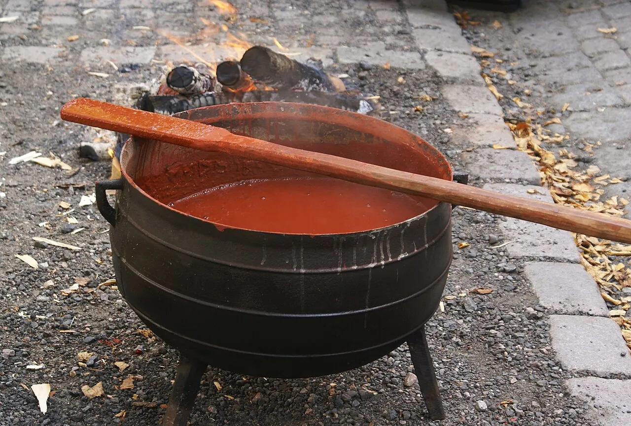 A black three-legged iron cauldron over a fire, full of bright red paint, with a long wooden paddle resting across it.