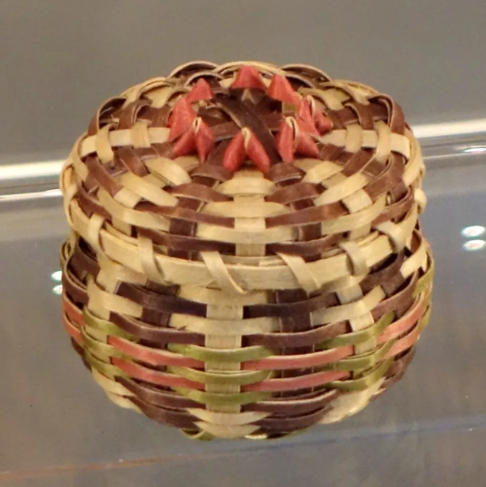 A small round lidded basket woven of natural, dark brown, and rose-dyed black ash splints, with curled points around the lid.